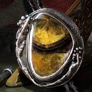 May include: A handcrafted pendant featuring a large, oval-shaped gemstone with yellow and brown bands, set in a textured silver bezel. The pendant has a unique, organic design, perfect for adding a touch of natural beauty to any outfit.