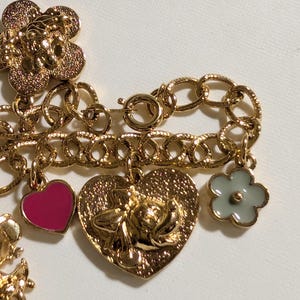May include: A gold-tone charm bracelet featuring a textured chain with multiple dangling charms, including a large gold flower, a small hot pink heart, a heart-shaped charm with a raised character face, and a small light gray flower charm.