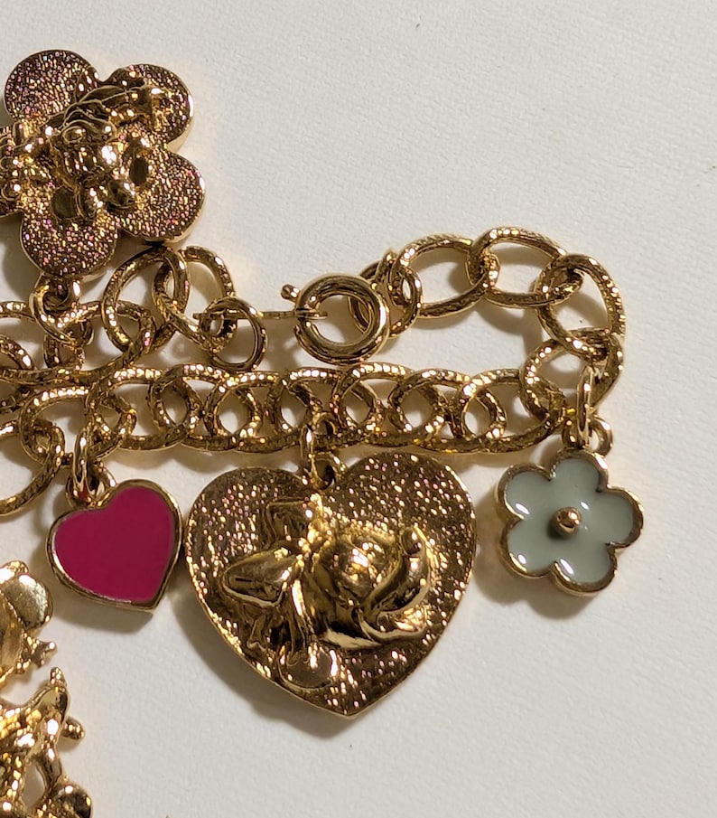 May include: A gold-tone charm bracelet featuring a textured chain with multiple dangling charms, including a large gold flower, a small hot pink heart, a heart-shaped charm with a raised character face, and a small light gray flower charm.