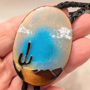 May include: An oval bolo tie featuring an enameled desert landscape scene. The design displays a bright blue sky, a dark saguaro cactus silhouette, and brown mountain shapes against a tan and orange ground. It is attached to a black braided cord.