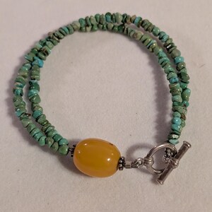May include: A double-strand bracelet featuring small turquoise beads and a large, oval amber-colored bead. The bracelet has a silver toggle clasp. The turquoise beads are a mix of green and blue.