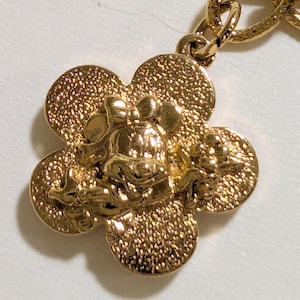 May include: A gold-toned metal charm featuring a textured, four-petaled flower shape with a raised, detailed depiction of a cartoon mouse character wearing a bow in the center. The charm is attached to a matching gold-toned metal chain link.