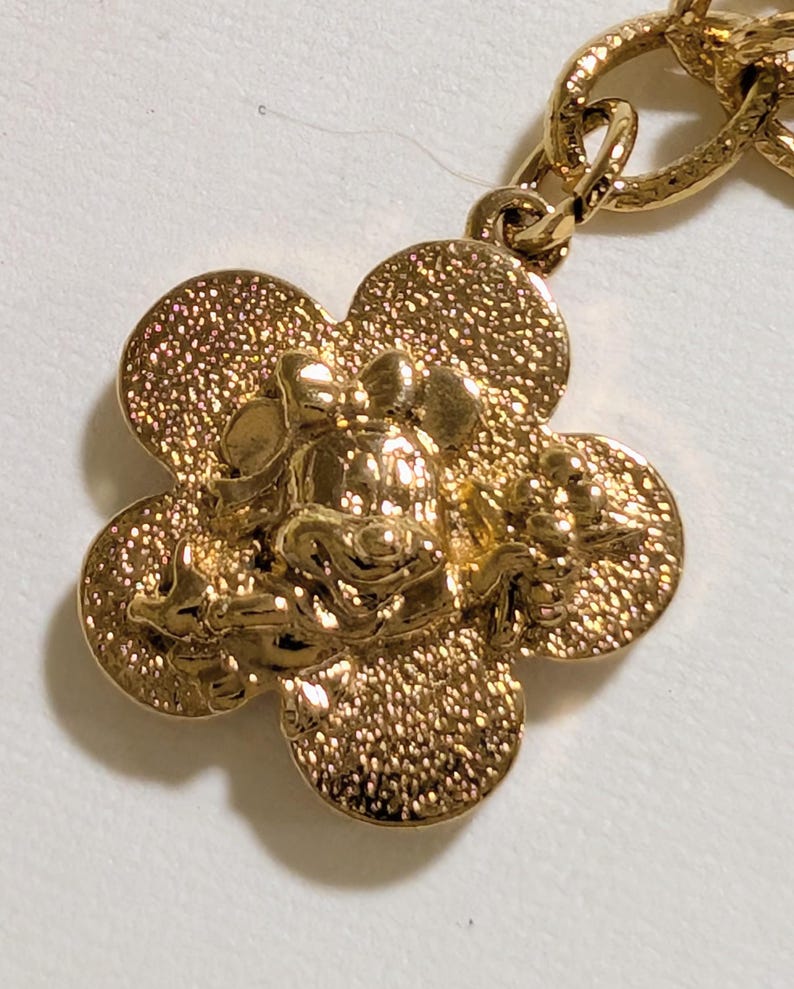 May include: A gold-toned metal charm featuring a textured, four-petaled flower shape with a raised, detailed depiction of a cartoon mouse character wearing a bow in the center. The charm is attached to a matching gold-toned metal chain link.