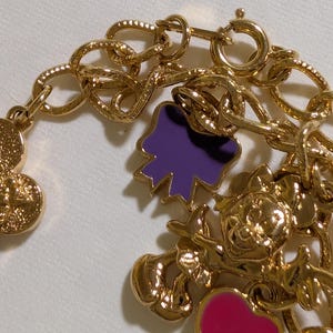 May include: A gold-tone charm bracelet featuring a series of metallic links and various dangling charms. The charms include a purple bow, a pink heart, and a detailed gold character figure, all attached to the chain with small circular jump rings.
