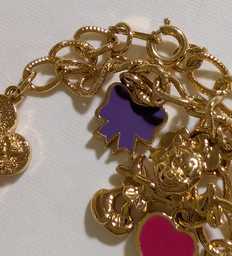 May include: A gold-tone charm bracelet featuring a series of metallic links and various dangling charms. The charms include a purple bow, a pink heart, and a detailed gold character figure, all attached to the chain with small circular jump rings.