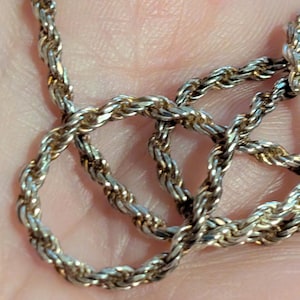 May include: Close-up of a silver-toned rope chain necklace. The chain is made of twisted metal links, creating a textured surface. The necklace is displayed against a blurred background.
