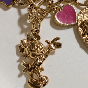 May include: A gold-tone charm bracelet featuring a polished gold-tone Minnie Mouse character charm, a small pink heart-shaped charm with enamel fill, and a purple enamel charm, all attached to a textured gold-tone chain link bracelet.