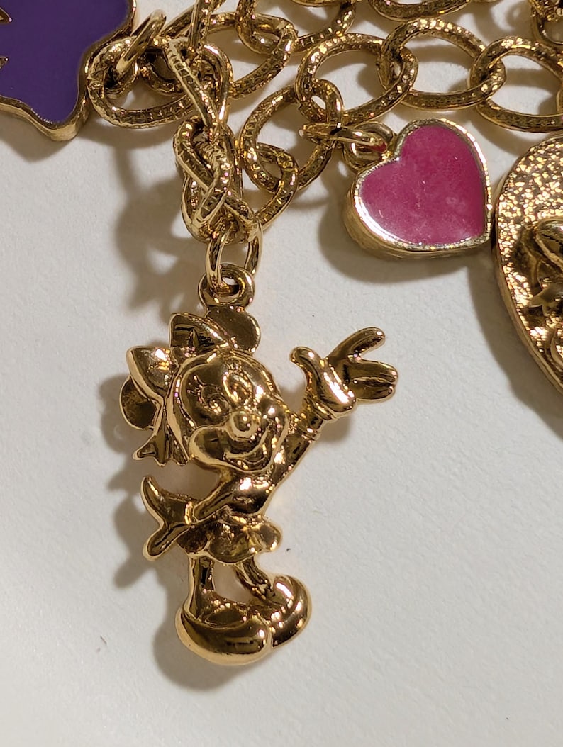 May include: A gold-tone charm bracelet featuring a polished gold-tone Minnie Mouse character charm, a small pink heart-shaped charm with enamel fill, and a purple enamel charm, all attached to a textured gold-tone chain link bracelet.