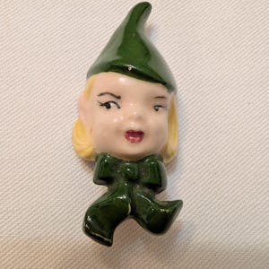 May include: A ceramic brooch shaped like a head with blonde hair, wearing a dark green pointed hat and a matching dark green scarf tied in a bow at the neck. The face features painted black eyebrows, eyelashes, and red lips on a glossy finish.