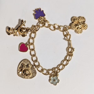 May include: A gold-tone charm bracelet featuring a textured chain link design. The bracelet includes several charms: a purple enamel bow, a gold-tone figure, a magenta enamel heart, a large textured gold heart, and a small light blue enamel flower.