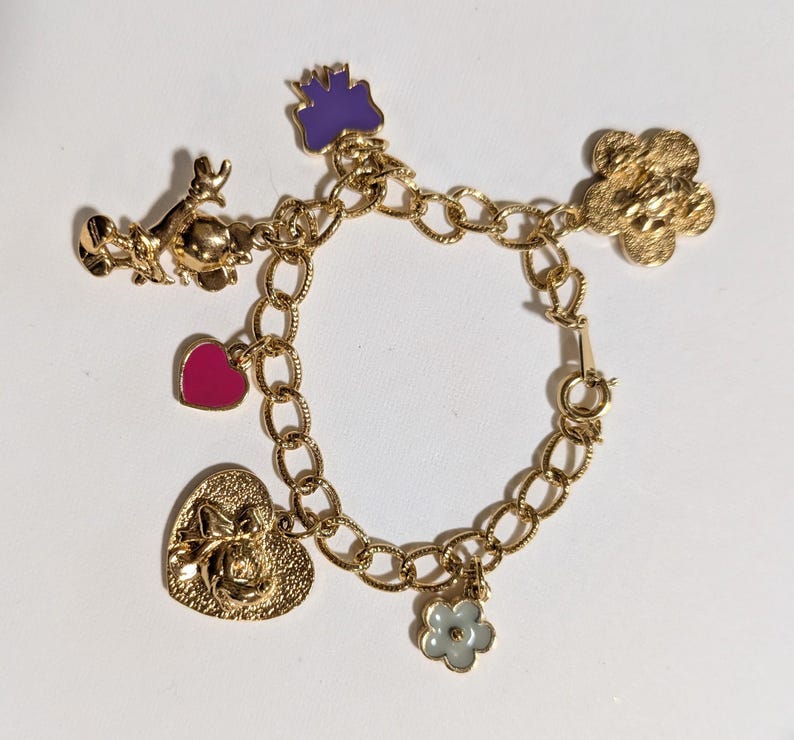 May include: A gold-tone charm bracelet featuring a textured chain link design. The bracelet includes several charms: a purple enamel bow, a gold-tone figure, a magenta enamel heart, a large textured gold heart, and a small light blue enamel flower.