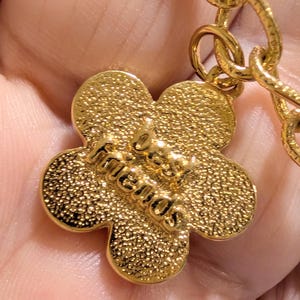 May include: A gold-toned metal charm shaped like a five-petaled flower with a textured surface. The words "best friends" are embossed in raised lettering across the center of the flower. The charm is attached to a gold-toned metal chain link.