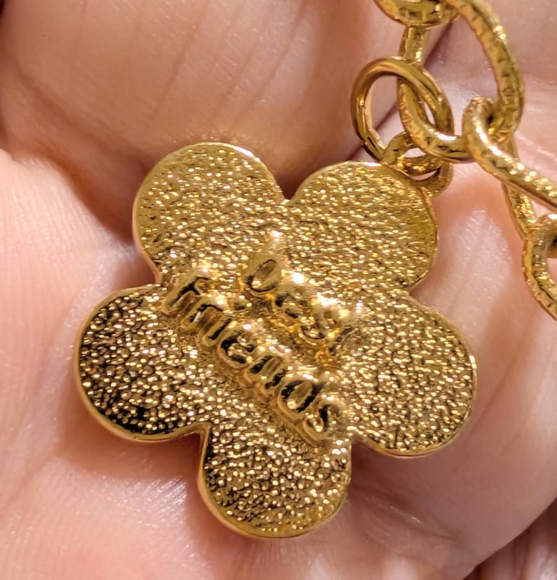 May include: A gold-toned metal charm shaped like a five-petaled flower with a textured surface. The words "best friends" are embossed in raised lettering across the center of the flower. The charm is attached to a gold-toned metal chain link.