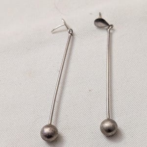 May include: A pair of silver-toned drop earrings featuring a long, thin metal rod suspended from a post back. Each earring terminates in a small, textured spherical bead at the bottom, creating a clean, linear design with a metallic finish.