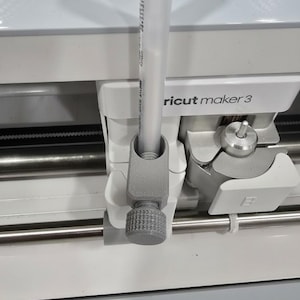May include: Close-up of a Cricut Maker 3 machine with a gray 3D-printed accessory holding a clear tube. The accessory has a textured knob. The machine is white and gray.