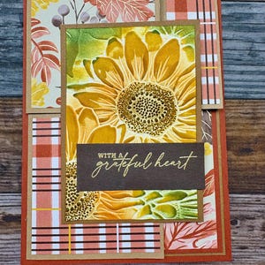 Può includere: Biglietto di auguri fatto a mano con un motivo a girasole. Il biglietto presenta un grande girasole in rilievo nei toni del giallo e del verde, con le parole "with a grateful heart" in lettere dorate. Lo sfondo include motivi a quadri e foglie.