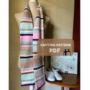 May include: A long, knitted scarf featuring horizontal stripes in shades of light blue, pink, white, tan, and dark purple. A rectangular overlay on the image contains the text KNITTING PATTERN PDF with a small icon of a person at a desk.