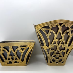 Peut inclure: Deux récipients avec un intérieur marron foncé et un extérieur doré présentant un motif complexe de volutes ajourées. Le plus petit récipient est large et peu profond, tandis que le plus grand est plus haut et s'évase vers le haut.