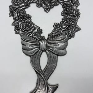 Può includere: Decorazione da parete a forma di cuore in metallo color argento. Il cuore è formato da rose e foglie, con un grande fiocco in basso. Il pezzo ha una superficie strutturata e dettagliata ed è probabilmente un oggetto decorativo.