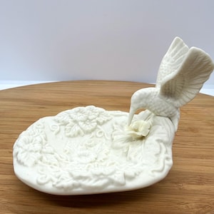 May include: A white ceramic hummingbird figurine perched above a floral-patterned dish. The hummingbird has outstretched wings and a long beak, appearing to feed from a flower. The dish is decorated with embossed flowers and leaves.
