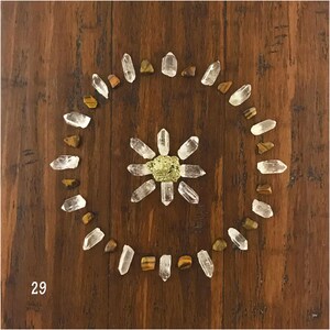 Puede incluir: Una rejilla de cristal con puntas de cuarzo transparente dispuestas en círculo alrededor de un cristal de pirita dorada sobre una superficie de madera marrón. El número 29 es visible en la esquina inferior izquierda.