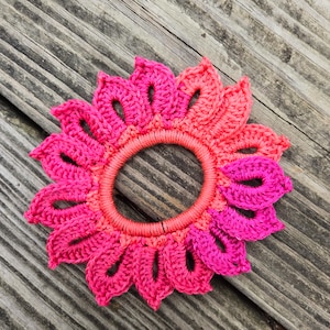 Puede incluir: Un coletero de ganchillo en forma de flor con pétalos rosas y coral.