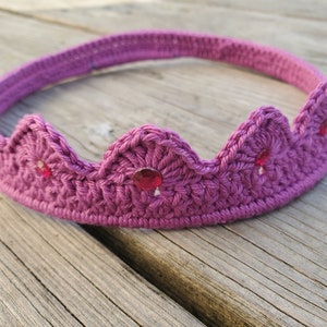 May include: A purple crocheted crown with red gem embellishments. The crown is made of yarn and has a simple design.