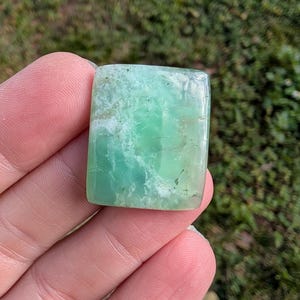 May include: A rectangular, polished gemstone with a translucent, light green hue. The stone has a slightly textured surface with white and darker green inclusions. The edges are rounded, and the overall shape is a smooth rectangle.