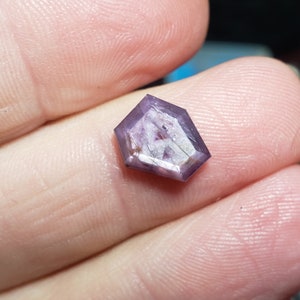 May include: A faceted purple gemstone with a hexagonal shape. The gemstone has a cloudy, translucent appearance with white and gray inclusions.