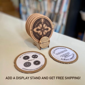 May include: Set of four cork coasters featuring a design of a manhole cover with the text "Copenhagen Kloakd&aelig;ksel" and "RethinkCopenhagen". The coasters are displayed on a wooden stand with the text "Manhole Coasters Copenhagen".