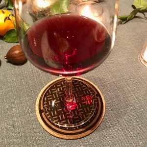 May include: A glass of red wine on a coaster with a black and white image of a New York City sewer cover. The coaster has the text "N.Y.C. SEWER" printed on it.