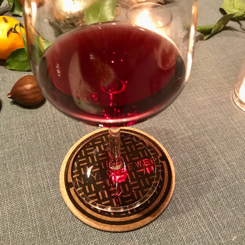 May include: A glass of red wine on a coaster with a black and white image of a New York City sewer cover. The coaster has the text "N.Y.C. SEWER" printed on it.