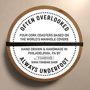 May include: A set of four cork coasters featuring designs based on manhole covers from around the world. The coasters are packaged in a round cardboard container with the text "Often Overlooked Always Underfoot" and the website address "www.tombino.shop".