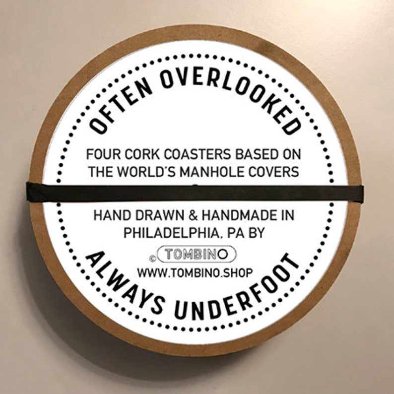 May include: A set of four cork coasters featuring designs based on manhole covers from around the world. The coasters are packaged in a round cardboard container with the text "Often Overlooked Always Underfoot" and the website address "www.tombino.shop".