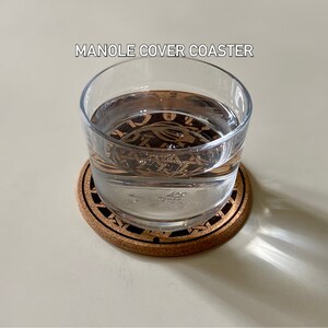 May include: A glass of water sits on a cork coaster with a black and brown design. The coaster has the text "MANOLE COVER COASTER" printed on it.
