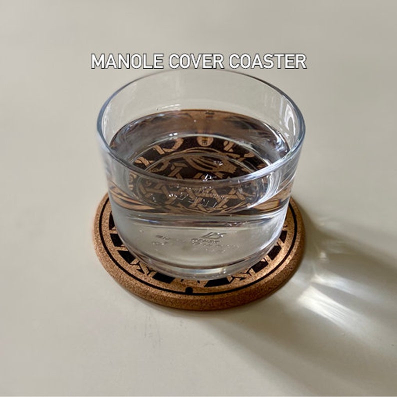 May include: A glass of water sits on a cork coaster with a black and brown design. The coaster has the text "MANOLE COVER COASTER" printed on it.