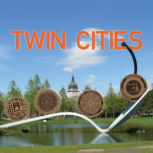 May include: A public art installation featuring the text "TWIN CITIES" in orange letters against a blue sky. The sculpture includes four circular medallions with city-related designs, suspended from a curved white structure over a pond.