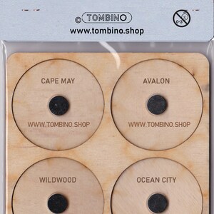 May include: Wooden plaque with four circular cutouts, each featuring a different New Jersey shore town name: Cape May, Avalon, Wildwood, and Ocean City. Each town name is printed in black text, and a small black circle is centered below the text. The text "WWW.TOMBINO.SHOP" is printed below each circle.