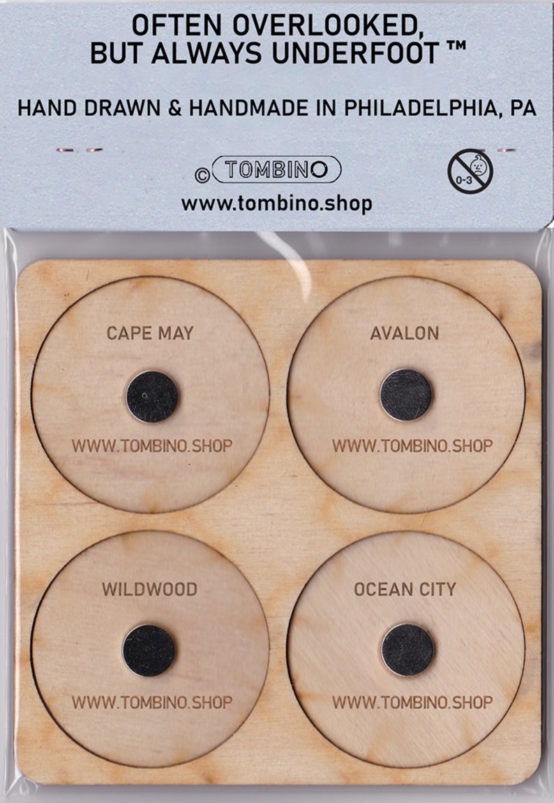 May include: Wooden plaque with four circular cutouts, each featuring a different New Jersey shore town name: Cape May, Avalon, Wildwood, and Ocean City. Each town name is printed in black text, and a small black circle is centered below the text. The text "WWW.TOMBINO.SHOP" is printed below each circle.