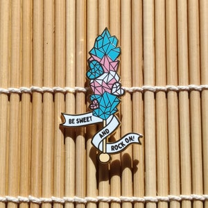 Op de afbeelding: Gouden pin met een blauw, roze en wit kristalontwerp. De pin heeft twee banners met de tekst "Be Sweet and Rock On!"