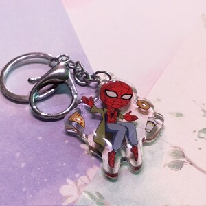 May include: A clear keychain featuring a cartoon illustration of Spider-Man sitting with a slice of pizza, a donut, and a cup of coffee. The keychain is attached to a silver metal ring.