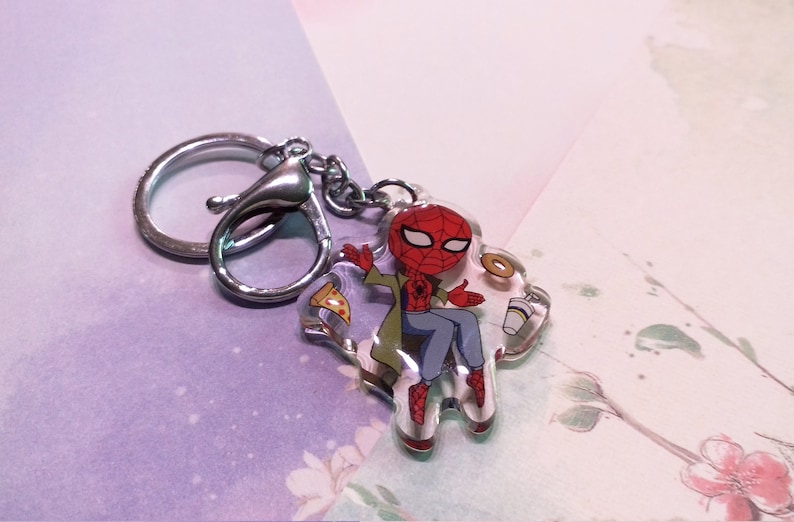 May include: A clear keychain featuring a cartoon illustration of Spider-Man sitting with a slice of pizza, a donut, and a cup of coffee. The keychain is attached to a silver metal ring.