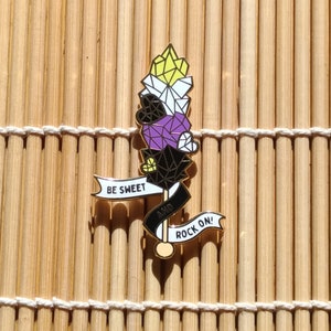 May include: Gold pin with a yellow, white, purple, and black geometric design. The pin has a banner that says "Be Sweet and Rock On!"