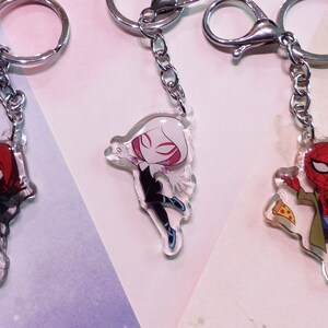 May include: Three different Spider-Man character keychains. The first keychain features Spider-Man Miles Morales in a red hooded suit. The second keychain features Spider-Gwen in a white and black suit. The third keychain features Spider-Man in a red and blue suit sitting with a slice of pizza and a cup of coffee.