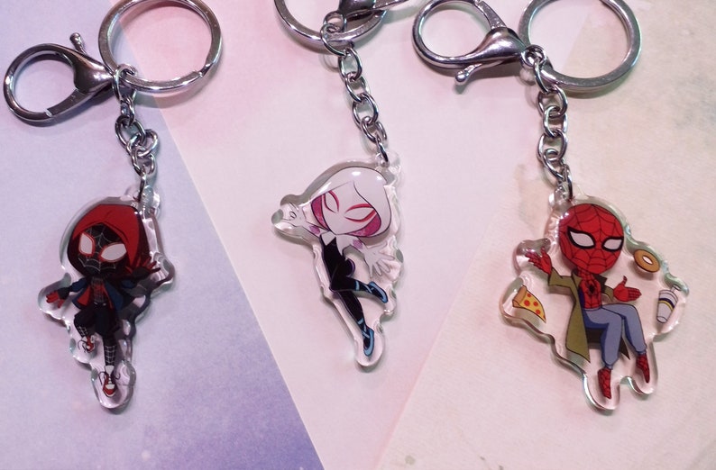May include: Three different Spider-Man character keychains. The first keychain features Spider-Man Miles Morales in a red hooded suit. The second keychain features Spider-Gwen in a white and black suit. The third keychain features Spider-Man in a red and blue suit sitting with a slice of pizza and a cup of coffee.