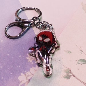 May include: A clear keychain with a red and black cartoon character wearing a red hooded jacket. The character is a stylized version of Spider-Man.