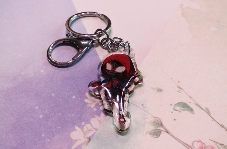 May include: A clear keychain with a red and black cartoon character wearing a red hooded jacket. The character is a stylized version of Spider-Man.