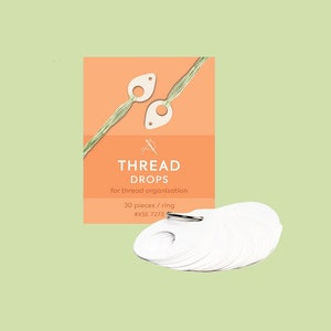 Könnte beinhalten: Ein Set mit 30 weißen Faden-Tropfen zur Organisation von Garn. Die Tropfen haben die Form einer Träne mit einem kleinen Loch oben. Die Tropfen sind in einer kleinen orangefarbenen Schachtel mit dem Text "THREAD DROPS for thread organisation 30 pieces / ring 8XSE_1212" verpackt.
