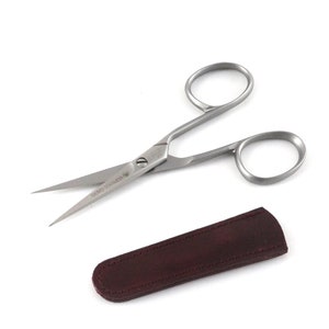 May include: Silver-toned small scissors with a burgundy leather case. The scissors have a curved handle and the words "DONO STAINLESS" are printed on the blade.