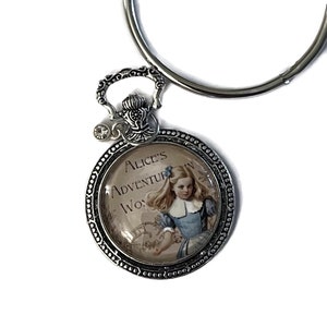 May include: Silver ring binder with a charm featuring a round glass cabochon with a vintage illustration of Alice from Alice's Adventures in Wonderland. The charm has a silver filigree design and a rhinestone.